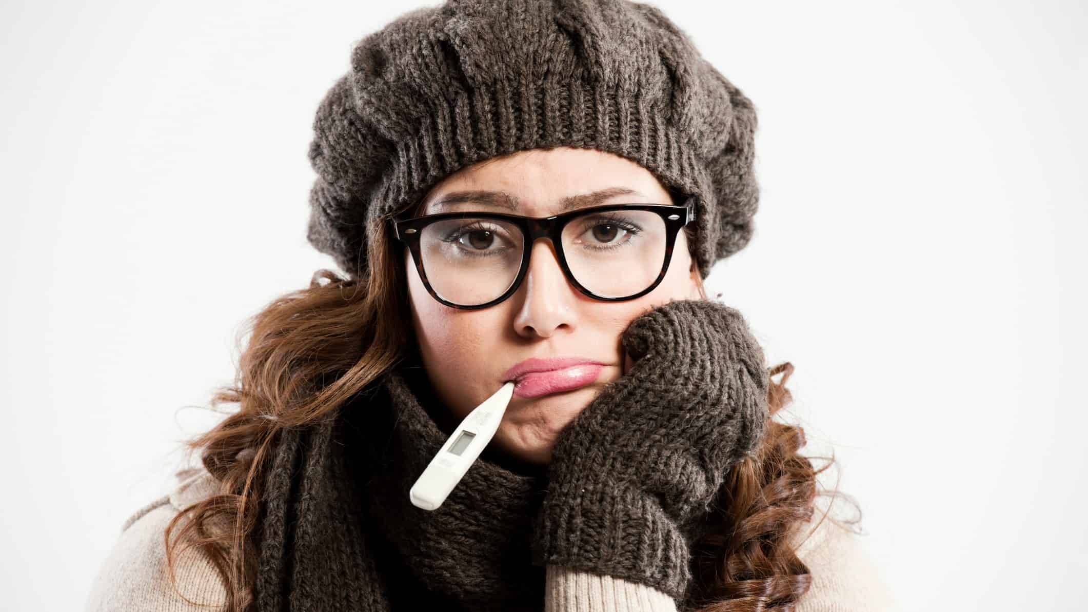 a woman rugged up in a woolen hat and gloves with a thermometer in her mouth props her hand under her chin as she looks dejectedly at the camera,, as though she is miserable from feeling sick.