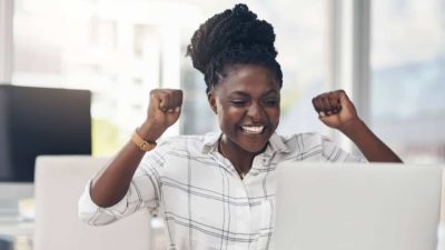 A young female ASX investor sits at her desk with her fists raised in excitement as she reads about rising ASX share prices on her laptop.