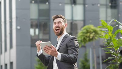 A male ASX investor on the street wearing a grey suit clenches his fist and yells yes after seeing on his ipad that the Paladin share price is going up again today