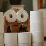 A young boy crouches behind a wall made of toilet rolls and uses two rolls as binoculars.