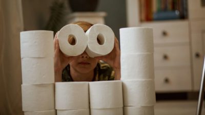 a young boy crouches behind a wall made of toilet rolls and uses two rolls as binoculars to hold up to his eyes as if guarding his stockpile.