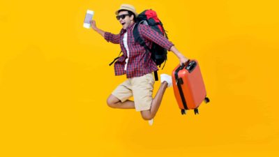 a tourist complete with suitcase and backpack with ticket in hand jumps for joy with his feet off the ground against a brightly coloured background.
