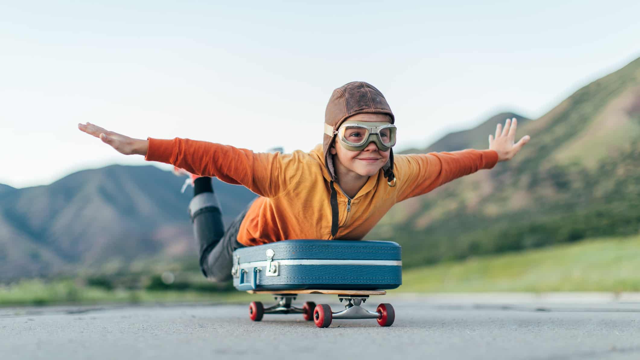 Kid with arm spread out on a luggage bag, riding a skateboard.