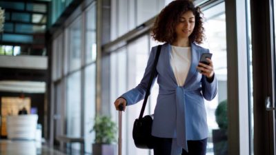 A corporate-looking woman looks at her mobile phone as she pulls along her suitcase in another hand while walking through an airport terminal with high glass panelled walls.