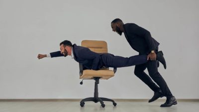 one man in a classic navy blue business suit lies atop a wheelie office shair while his colleage, also in a navy business suit, grabs him by the legs and propels him forward with both of them smiling widely as though larking about in the office.