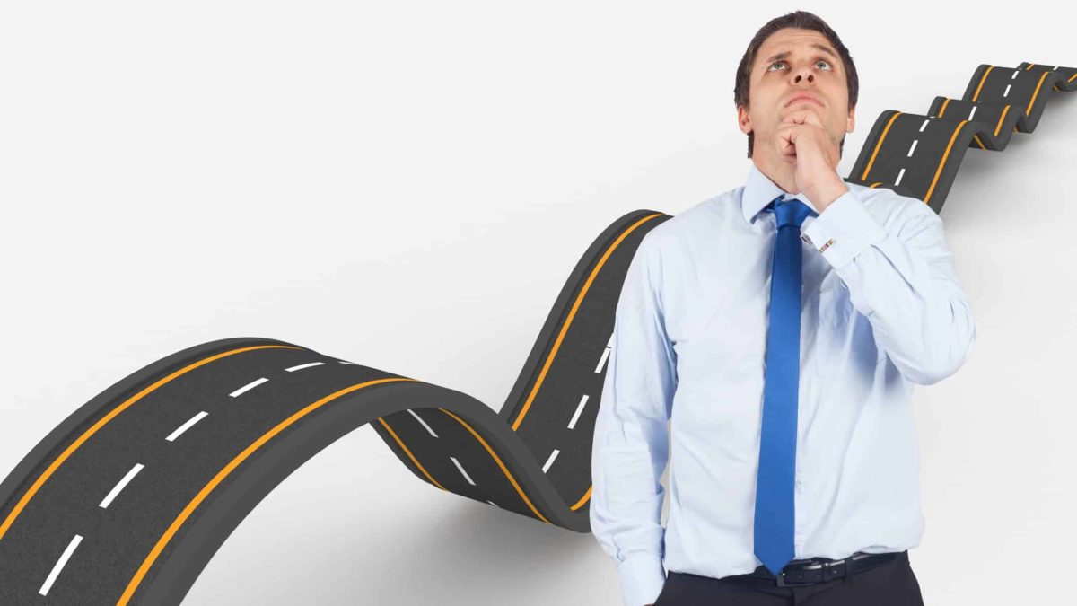 a man in a shirt and tie holds his chin in thoughtful contemplation and looks skywards as if thinking about something while a graphic of a road with many ups and downs unfurls behind him.