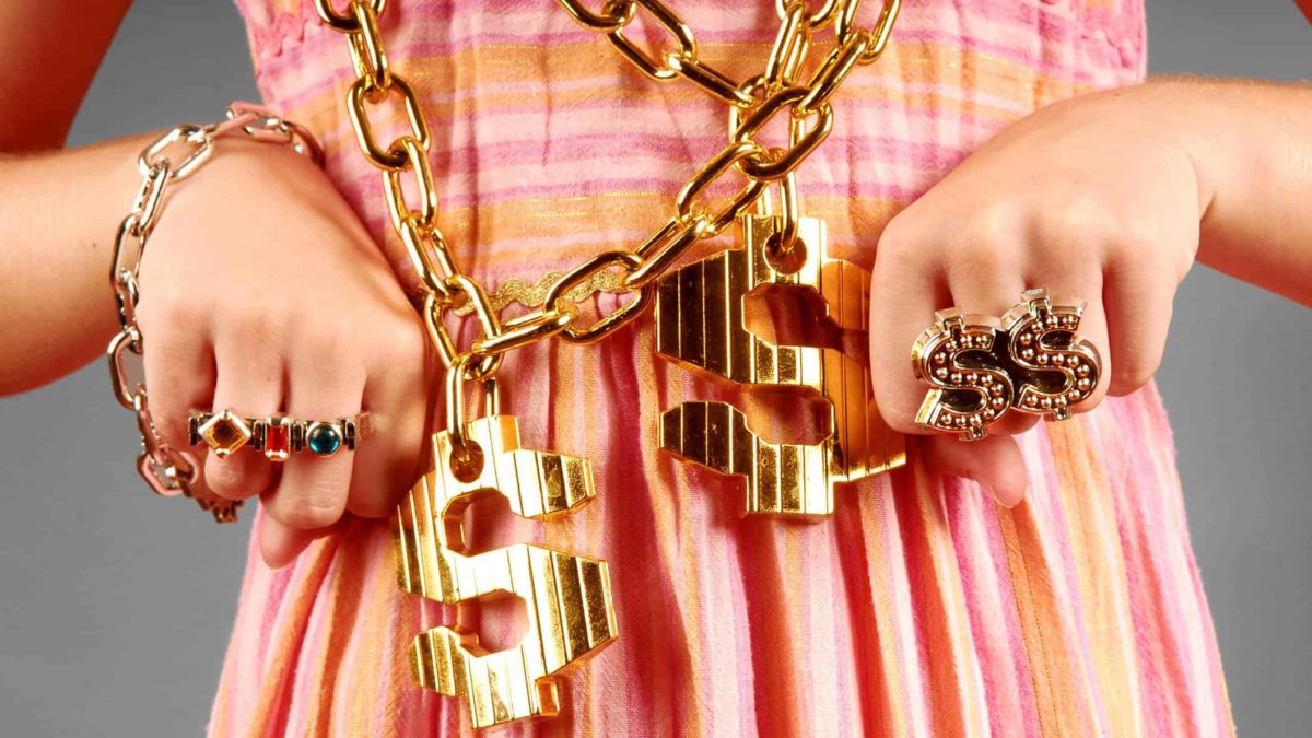 A young woman's hands are shown close up with many blingy gold rings on her fingers and two large gold chains around her neck with dollar signs on them.