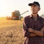 a wheat farmer stands with his arms crossed in a paddock of wheat ready for harvest with his header harvesting equipment operating in the background.