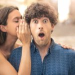 A man looks surprised as a woman whispers in his ear.
