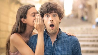 A man looks surprised as a woman whispers in his ear.