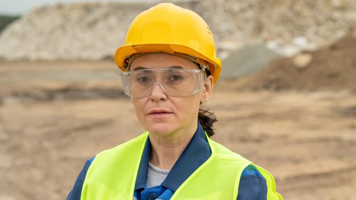 a female miner looks straight ahead at the camera wearing a hard hat, protective goggles and a high visibility vest standing in from of a mine site and looking seriously with direct eye contact.