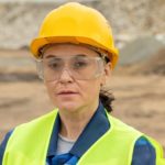 a female miner looks straight ahead at the camera wearing a hard hat, protective goggles and a high visibility vest standing in from of a mine site and looking seriously with direct eye contact.