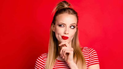 A young woman wearing a red and white striped t-shirt puts her hand to her chin and looks sideways as she wonders whether to buy NAB shares