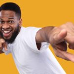 A young bearded man wearing a white t-shirt with a yellow backdrop holds up his arms to his chest and points to the camera in celebration of ASX shares rising today