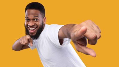 A young bearded man wearing a white t-shirt with a yellow backdrop holds up his arms to his chest and points to the camera in celebration of ASX shares rising today