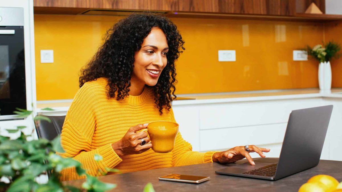 A woman wearing yellow smiles and drinks coffee while on laptop.