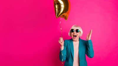 A cool older woman wearing sunglasses celebrates at her party with a gold balloon.