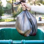 A hand holds a garbage bag over a wheelie bin, about to dump the rubbish.
