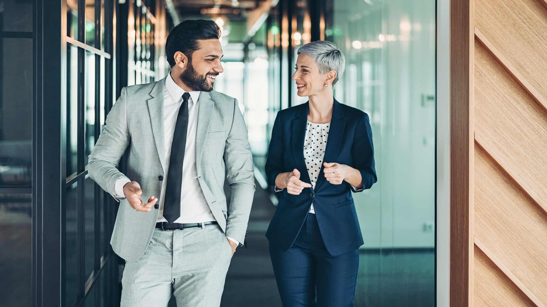 Two businesspeople walk together in an office, smiling as they enjoy a good business relationship.