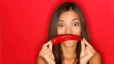 A young woman holds a red chilli in front of her mouth with eyes wide open looking happy about the Hot Chili share price today