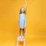 A little girl stands on a chair and reaches really, really high with her hand, in front of a yellow background.