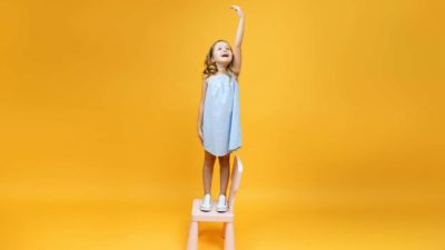 A little girl stands on a chair and reaches really, really high with her hand, in front of a yellow background.