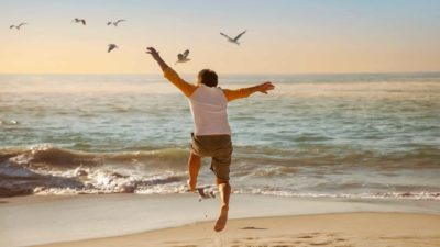 A boy leaps and flaps his arms as he tries to fly with some birds on the shoreline of the beach.