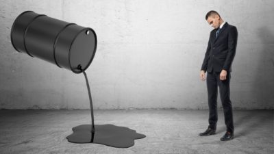 A barrel of oil suspended in the air is pouring oil while a man in a suit stands with a droopy head watching the oil drop out.