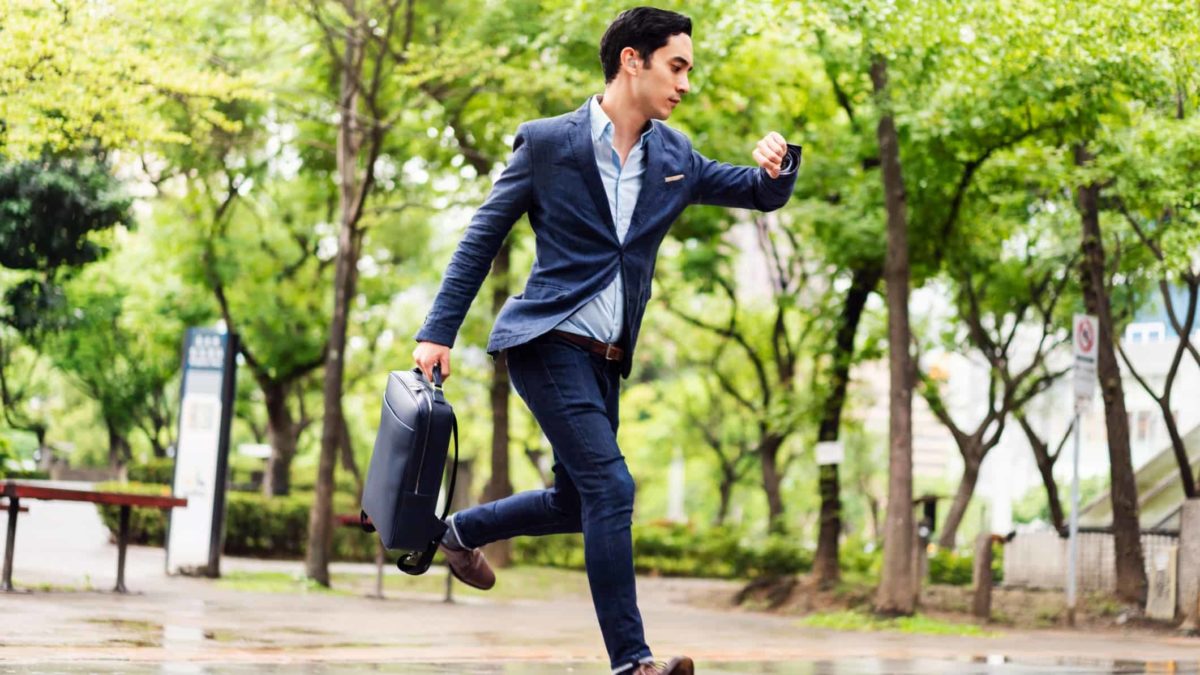 A man wearing a suit and holding a briefcase looks at his watch as he runs across a park, running late.