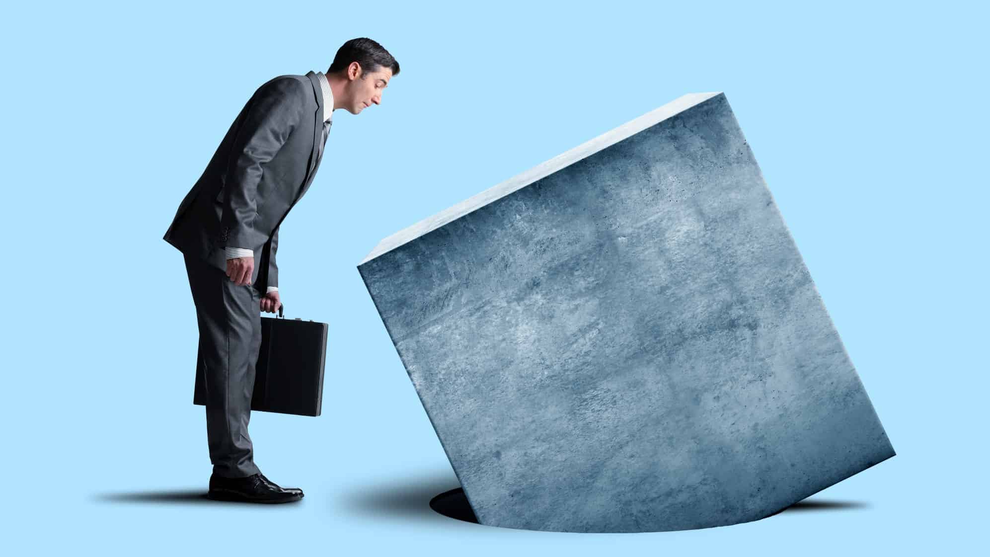 A businessman carrying a briefcase looks at a square peg or block sinking into a round hole.