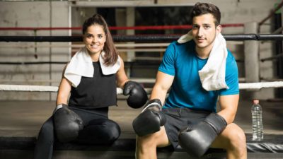 a man and a woman sit on the edge of a boxing ring wearing boxing gloves and with towels around their shoulders as they smile, as if they have just finished a boxing workout.