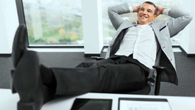 a man in a business suit sits happily leaning back into his hands behind his head with his feet on his desk and smiles broadly.