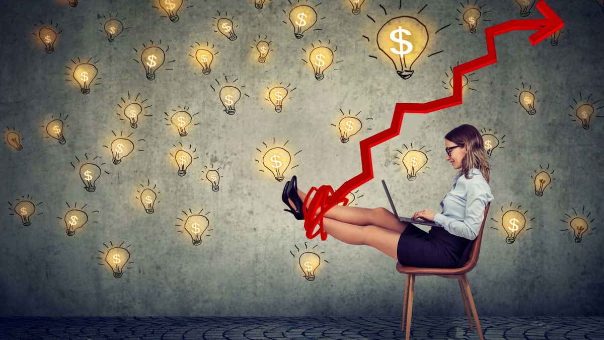 A woman sits on a chair with laptop on her lap and a smile on her face with a graphic image of a climbing jagged arrow tangled around her feet and lifting them comfortably so they are raised against a backdrop of many lightbulbs with one large lightbulb showing a dollar sign.