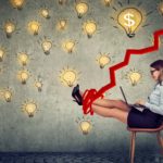 A woman sits on a chair with laptop on her lap and a smile on her face with a graphic image of a climbing jagged arrow tangled around her feet and lifting them comfortably so they are raised against a backdrop of many lightbulbs with one large lightbulb showing a dollar sign.