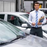 a car dealer stands amid a selection of cars parked in a showroom while he is holding a set of keys and paperwork in his other hand.