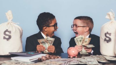 two young boys dressed in business suits and wearing spectacles look at each other in rapture with wide open mouths and holding large fans of banknotes with other banknotes, coins and a piggybank on the table in front of them and a bag of cash at the side.