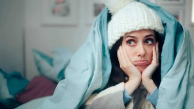 A young woman looking cold and bored rugged up and staying under the covers while the electricity is out representing Strike Energy shares in a trading halt today