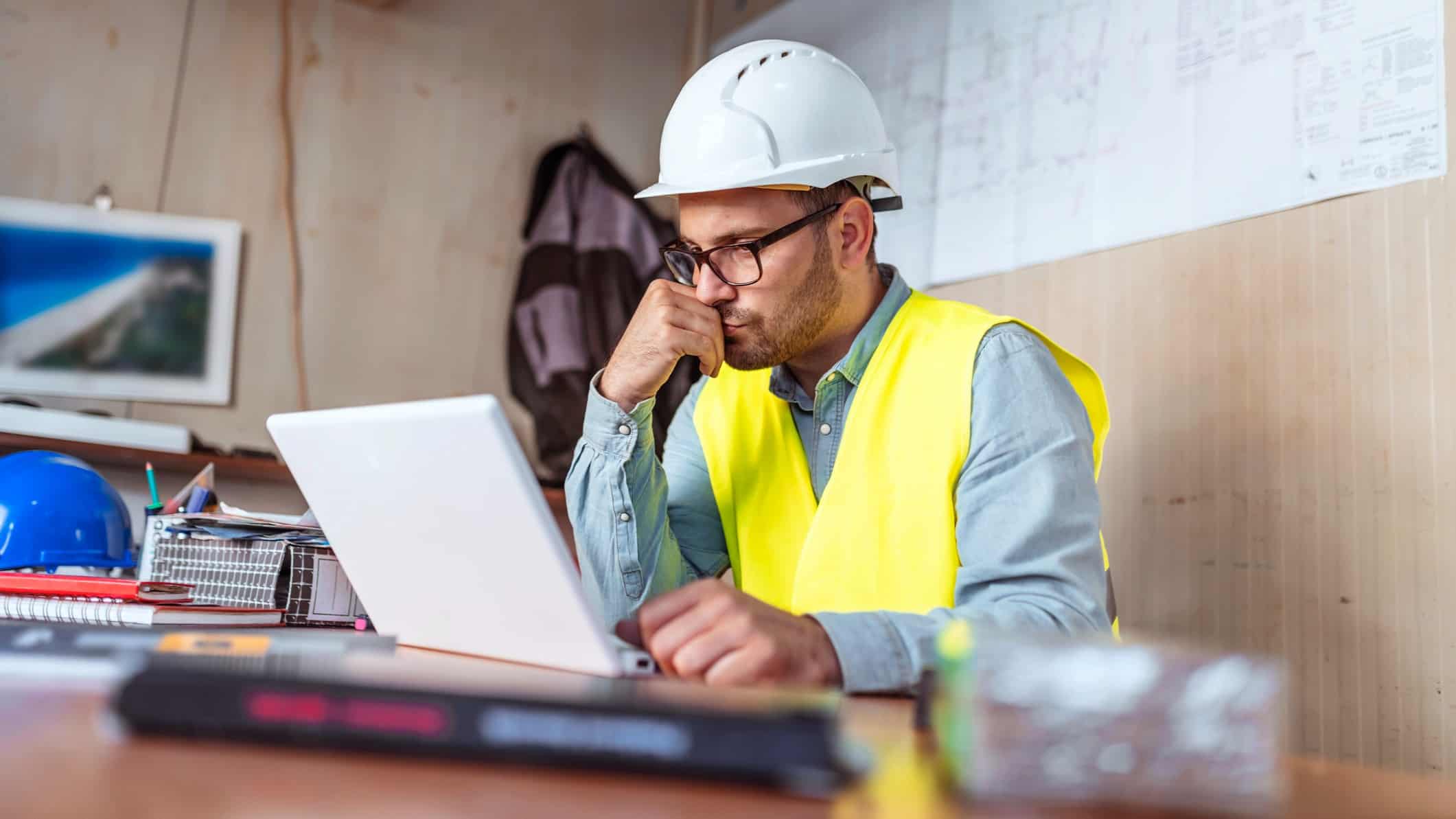 a construction worker sits pensively at his desk with his arm propping up his chin as he looks at his laptop computer while wearing a hard hat and visibility vest in a bunker style construction shed.