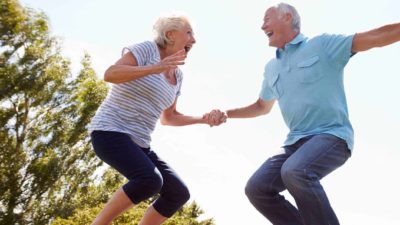 An older couple holding hands as they laugh while bouncing on a trampoline feeling happy about earning dividends from their ASX shares.
