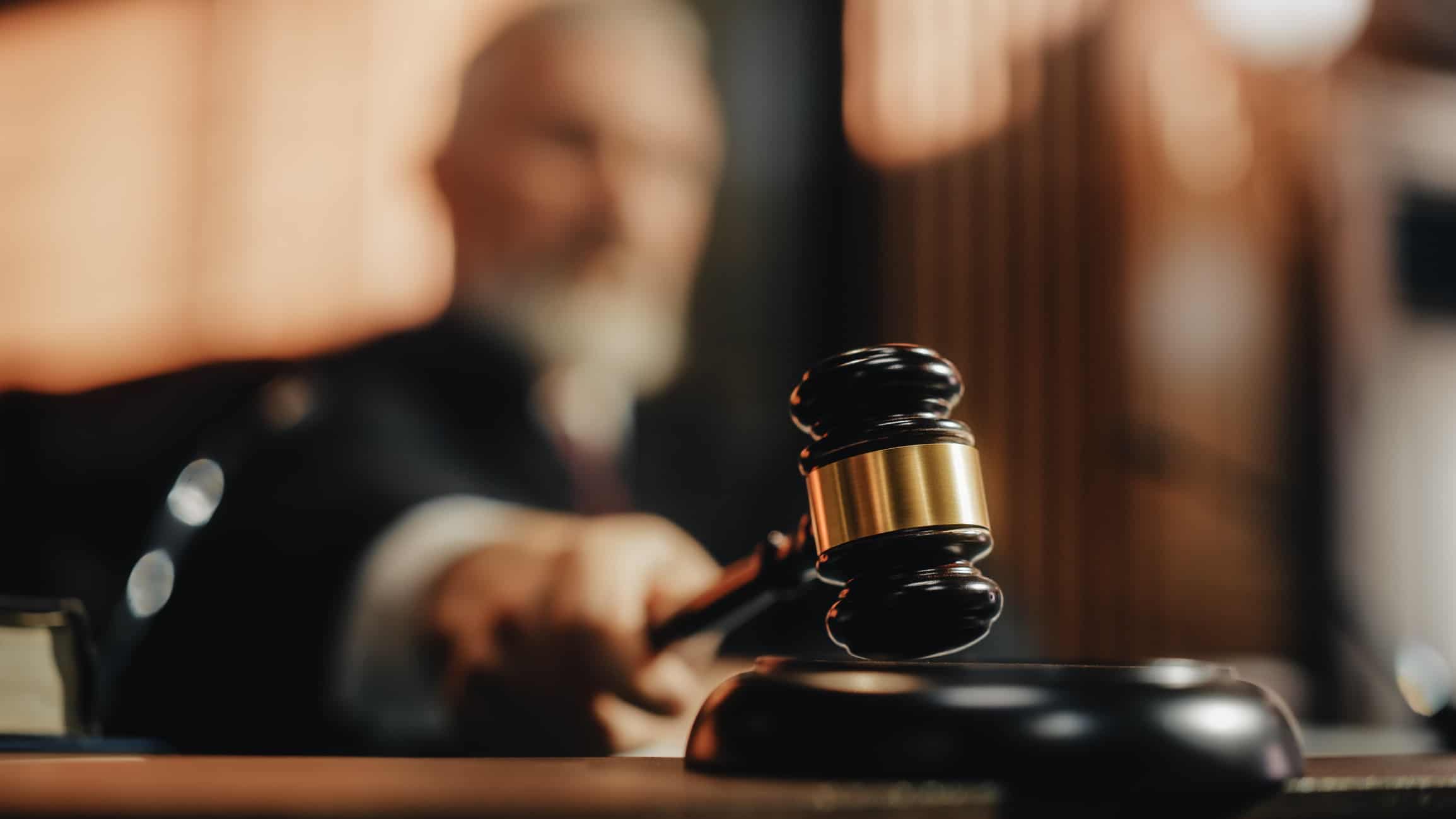 a judge sitting in a blurred background reaches forward to strike his gavel on the strikeplate on his judge's bench.