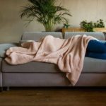 A young woman lies on her lounge with a pink blanket covering her face and the top half of her body as she hides away from seeing the Nick Scali share price fall today