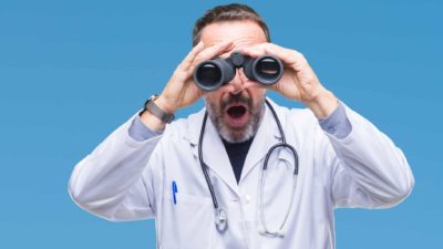A doctor appears shocked as he looks through binoculars on a blue background.
