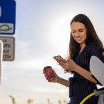 a woman smiles as she checks her phone in one hand with a takeaway coffee in the other as she charges her electric vehicle at a charging station.
