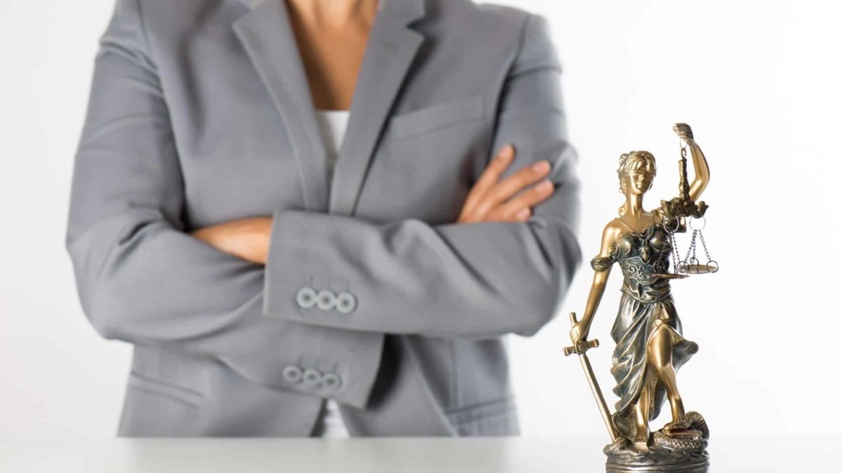a woman in a business suit stands with her arms folded in the background of a statue of lady justice wearing robes, carrying a sword and holding the scales of justice.