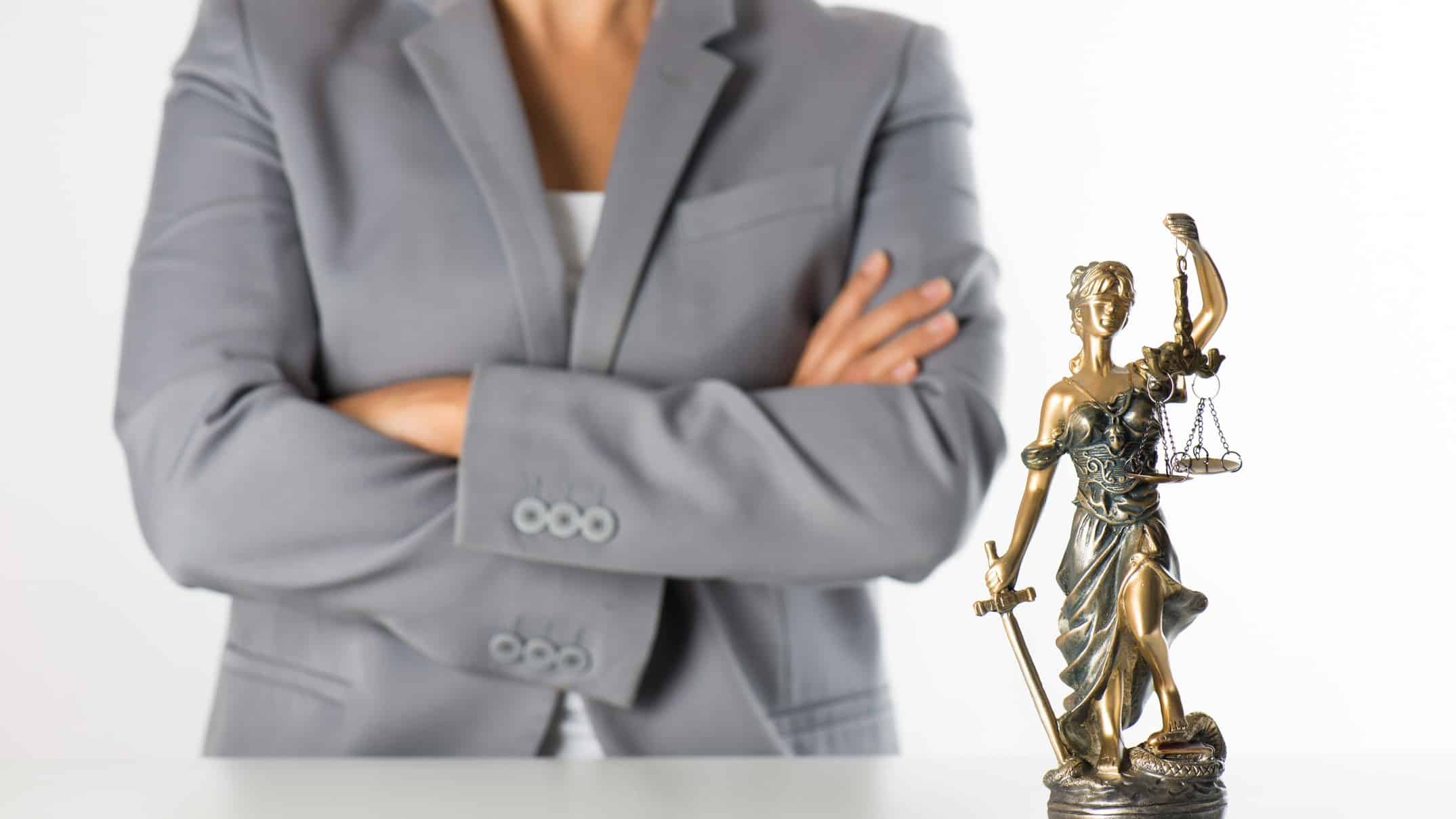 a woman in a business suit stands with her arms folded in the background of a statue of lady justice wearing robes, carrying a sword and holding the scales of justice.