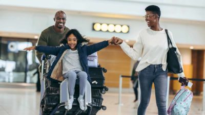 Two adults and a child look happy as they walk through airport with child sitting on suitcase.