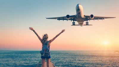 A woman reaches her arms to the sky as a plane flies overhead at sunset.