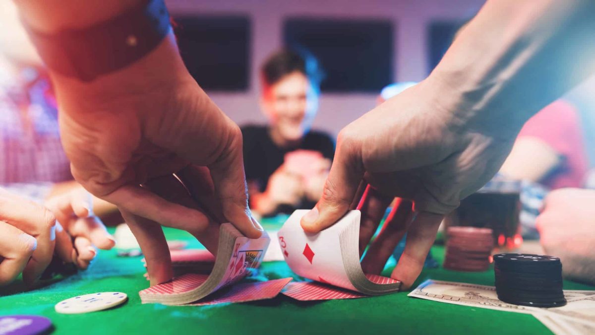 a close up of a casino card dealer's hands shuffling a deck of cards at a professional gambling table with the eager faces of casino patrons in the background.