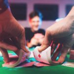 a close up of a casino card dealer's hands shuffling a deck of cards at a professional gambling table with the eager faces of casino patrons in the background.