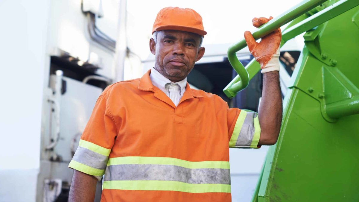 a man wearing a high visibility vest and safety gloves holds onto the back of a green garbage truck and looks straight at the camera with a workmanlike expression on his face.